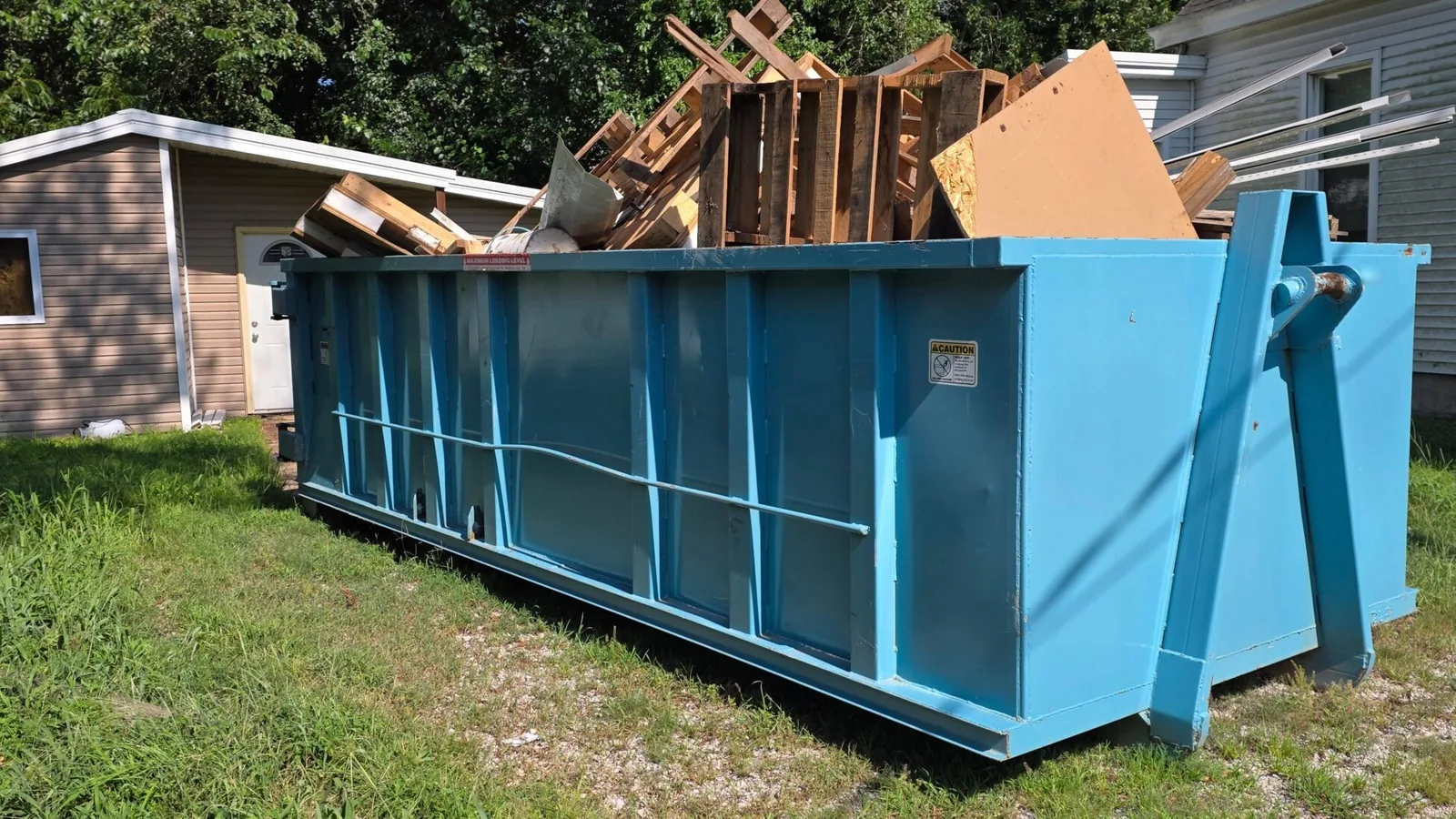 Blue roll-off dumpster loaded with pallets and remodel scrap beside a house and a shed, 30 yard dumpster rental