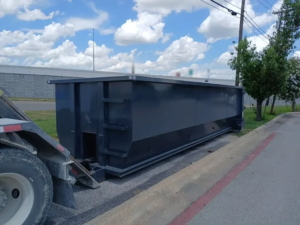 Large dark gray roll-off dumpster beside a curb and a highway sound wall, commercial dumpster rental