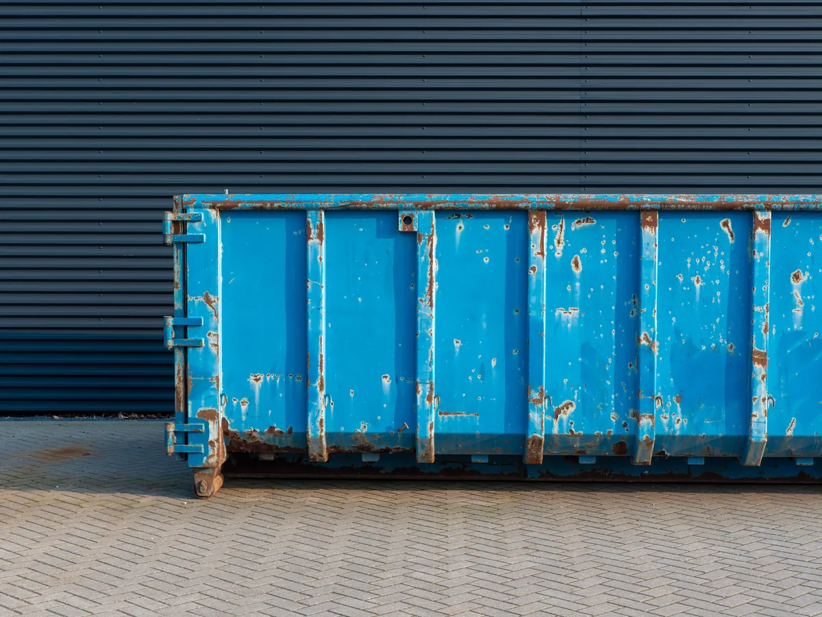 Weathered blue roll-off dumpster against a dark metal warehouse wall, commercial dumpster rental