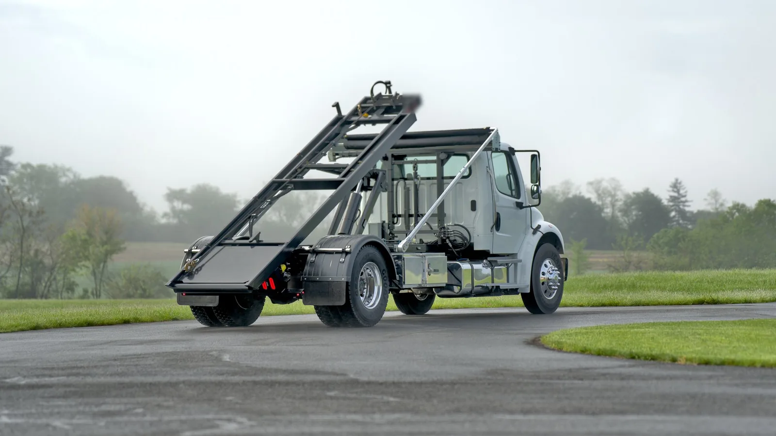 Roll-off truck with its hoist rails raised on a paved lot beside a field, roll-off dumpster rental