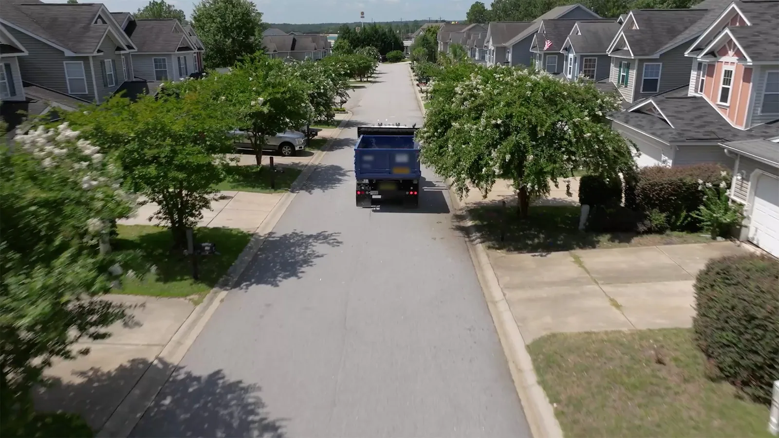 Roll-off truck carrying a blue roll-off dumpster down a tree-lined residential street, dumpster rental service area