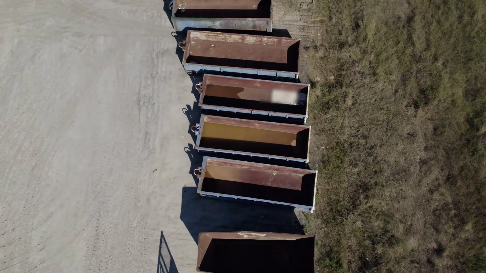 Aerial view of a row of empty roll-off dumpsters on a gravel lot beside dry grass, dumpster rental services