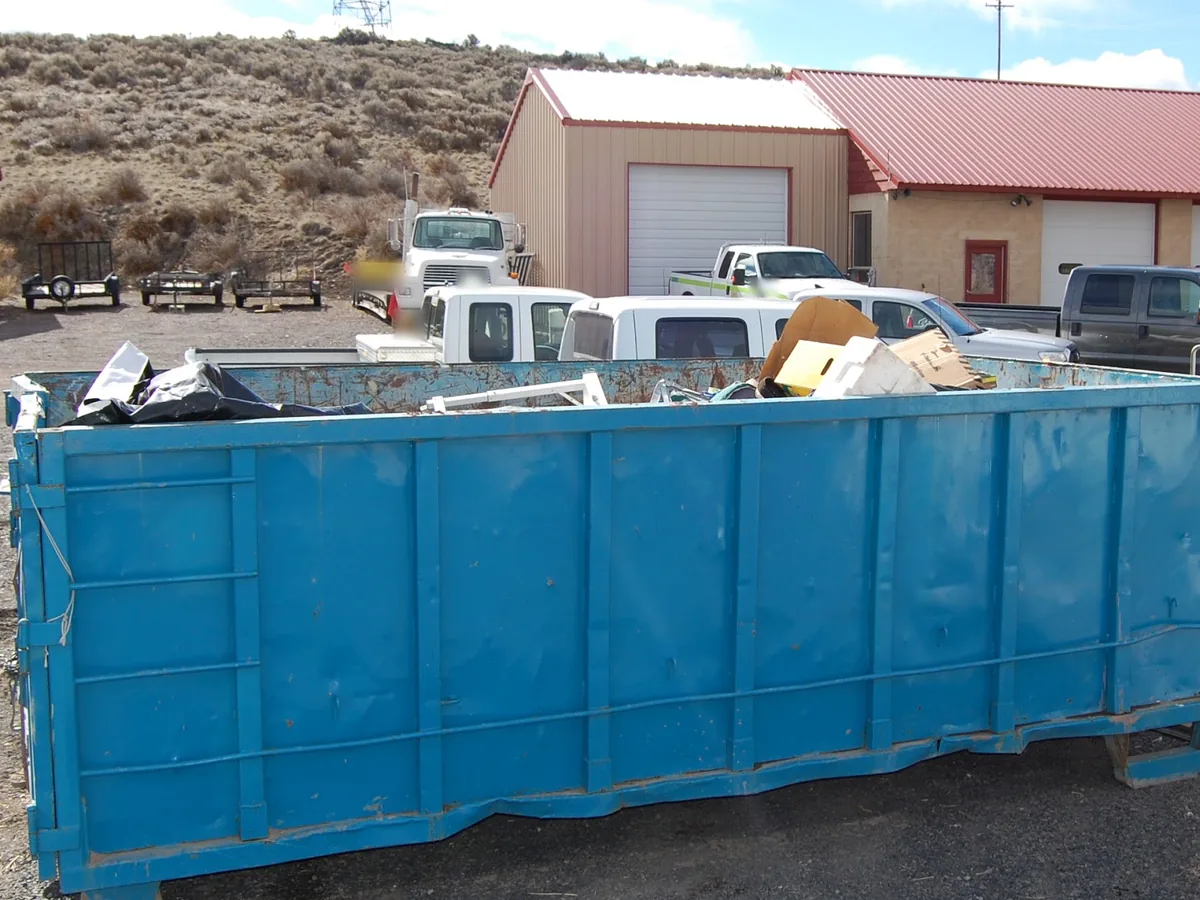 Teal roll-off dumpster loaded with cardboard and bagged trash beside a gravel lot, residential dumpster rental