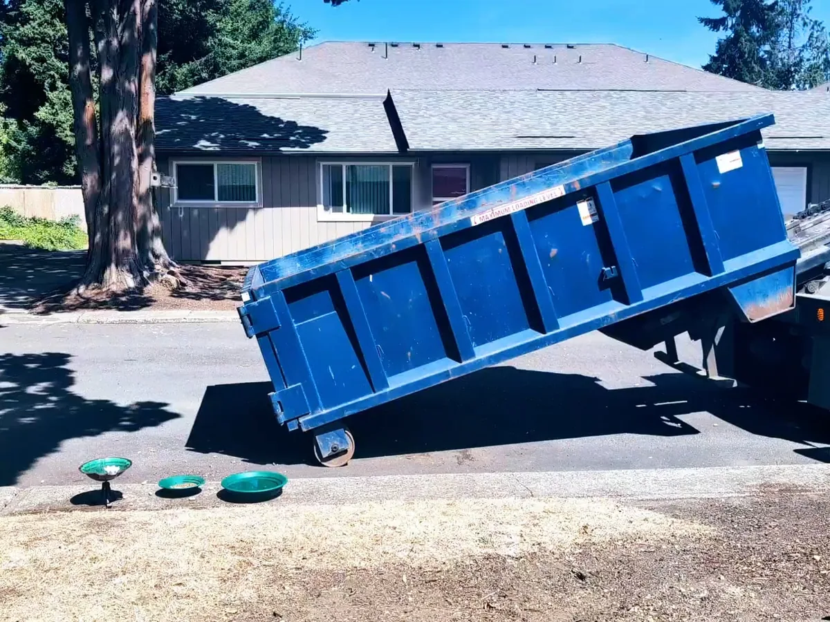 Blue roll-off dumpster tilted on the truck rails as it is set down in front of a house, roll-off dumpster rental