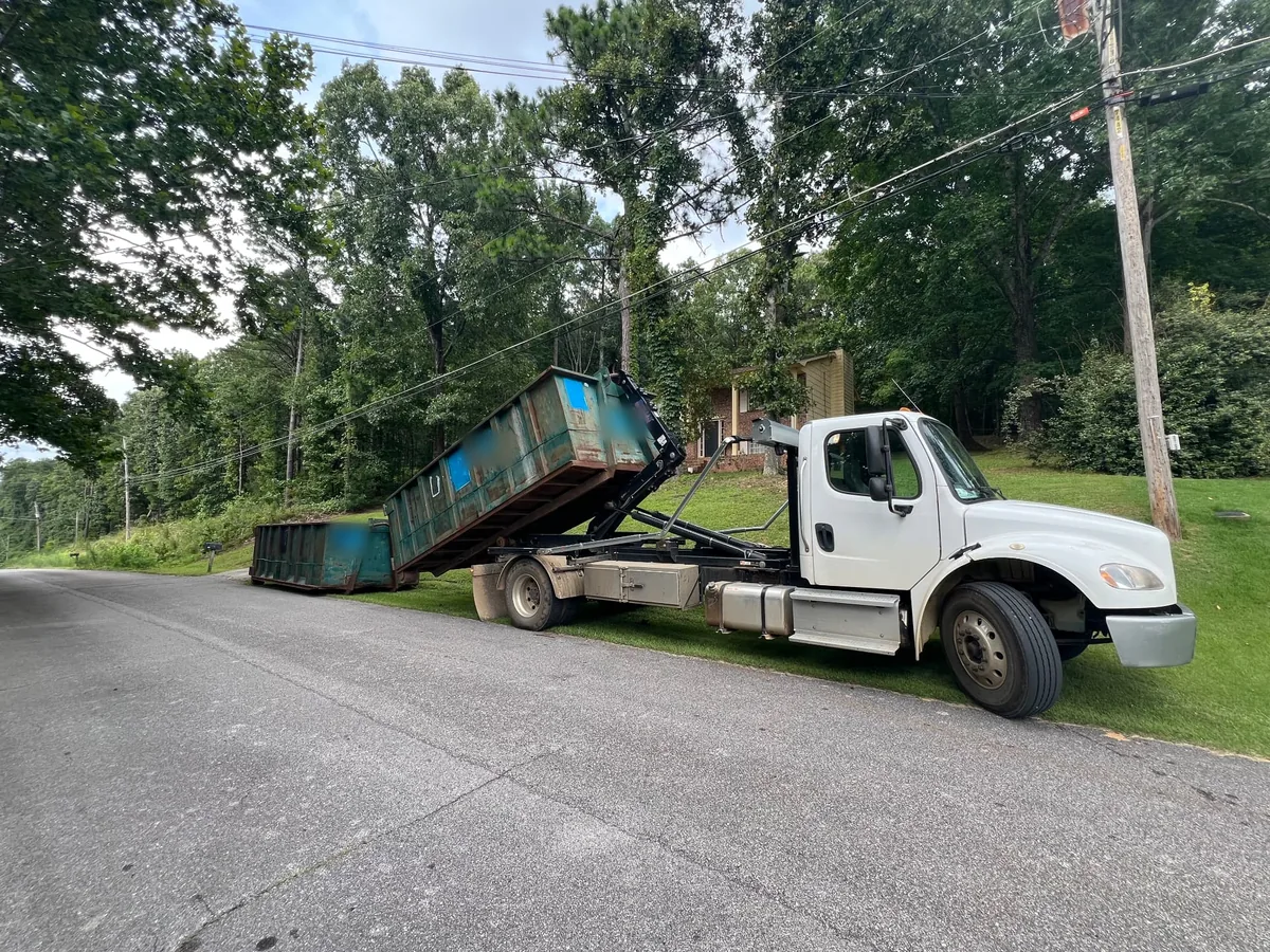 Roll-off truck lifting a blue-green roll-off dumpster onto its rails on a wooded road, roll-off dumpster rental