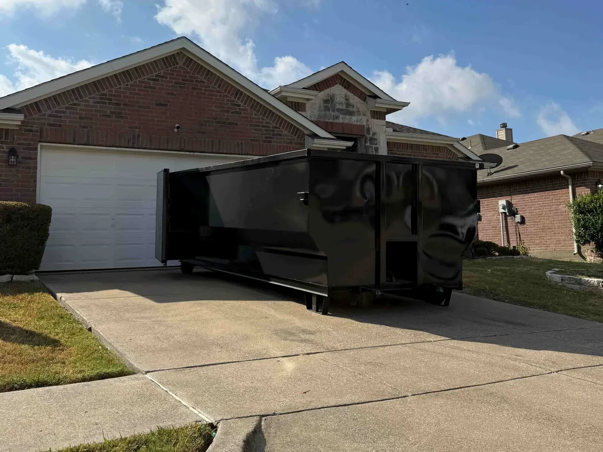 Black roll-off dumpster on the concrete driveway of a brick home, same-day dumpster rental